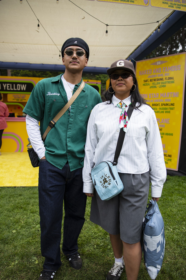A man and woman stand side by side outdoors; the man wears a green shirt and sunglasses, the woman wears a striped shirt, cap, sunglasses, and carries bags.